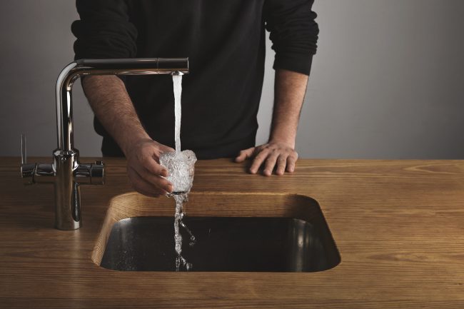 Barista rinses small glass with water Brutal barista in black sweatshot behind thick wooden table rinses small tranparent glass with water under silver metal tap in cafe shop. Water drops out of glass.